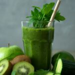 Vibrant emerald green detox smoothie in a glass with straw garnished with fresh mint, cucumber, and green apple
