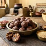 Cluster of glossy dark brown Simili Urundai ragi sesame peanut ladoos in an antique brass uruli with cracked open ball showing crushed peanuts